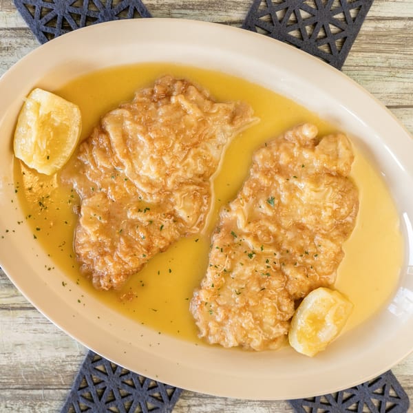 Two pieces of chicken piccata in a lemon-butter sauce with lemon wedges on a white plate.