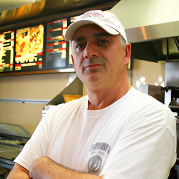 Romano's Pizza owner, a man in a white cap and t-shirt, stands in his kitchen.