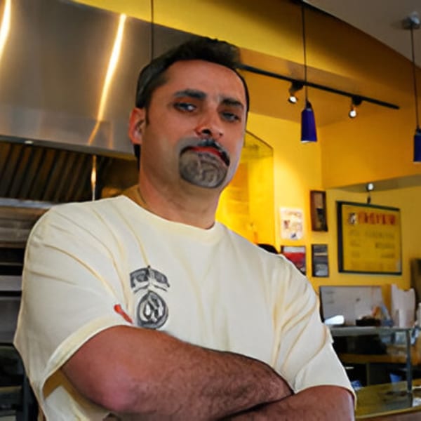 Man with goatee and mustache, wearing a light yellow shirt, standing in a pizza kitchen.